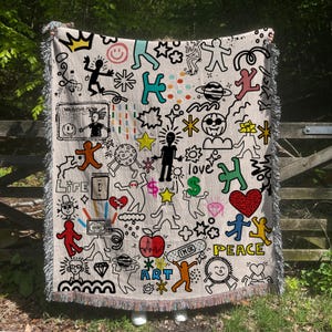 Può includere: Coperta beige con un colorato design a scarabocchi. La coperta presenta varie illustrazioni, tra cui omini stilizzati, stelle, cuori e le parole "love", "art" e "peace". La coperta ha un bordo sfrangiato.