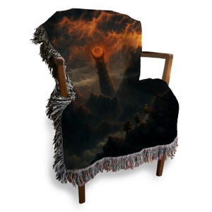 Puede incluir: Una manta tejida sobre una silla de madera. La manta presenta un paisaje oscuro y dram&aacute;tico con un cielo ardiente y una torre alta y ominosa. Los bordes de la manta tienen un ribete con flecos de varios colores.