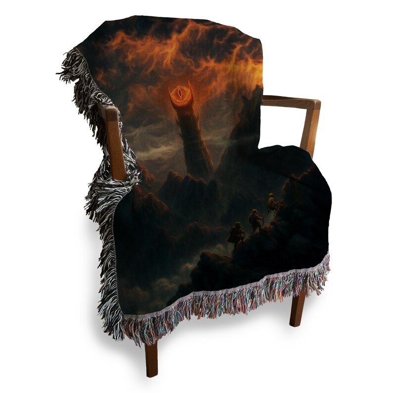 Puede incluir: Una manta tejida sobre una silla de madera. La manta presenta un paisaje oscuro y dram&aacute;tico con un cielo ardiente y una torre alta y ominosa. Los bordes de la manta tienen un ribete con flecos de varios colores.