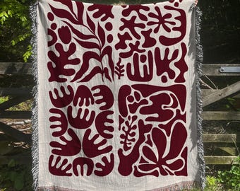 May include: A woven throw blanket with a burgundy abstract pattern of leaves and flowers on a cream background. The blanket has fringe on all sides.