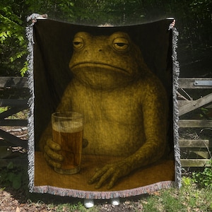 May include: Woven throw blanket featuring a humorous illustration of a frog holding a beer. The frog is depicted with a weary expression, seated at a table. The blanket has a fringed edge and is primarily in shades of brown and green.