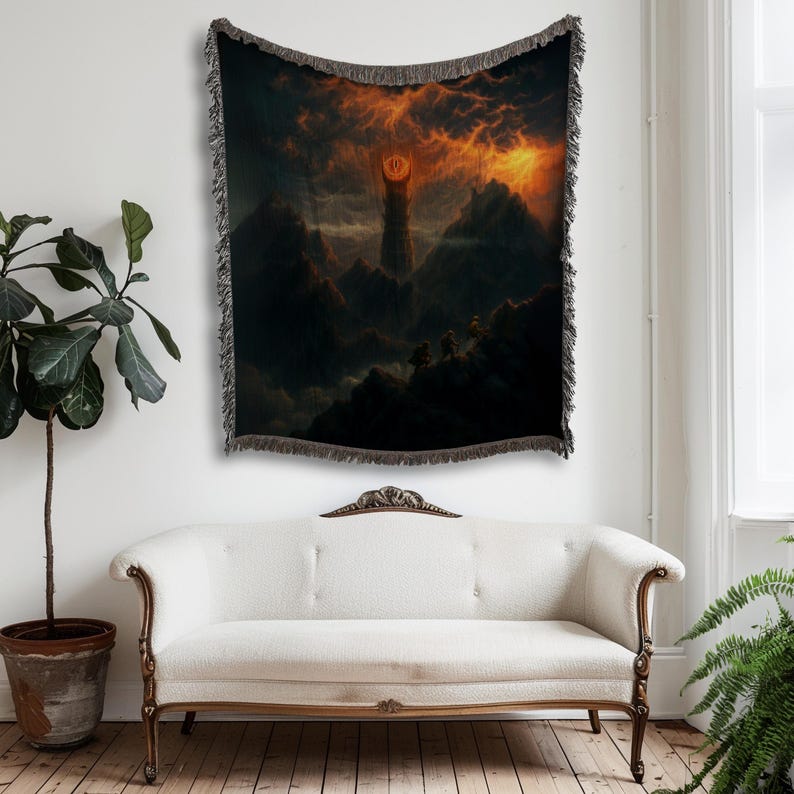 Puede incluir: Un tapiz tejido que presenta un paisaje dram&aacute;tico con una alta torre bajo un cielo de fuego. La obra de arte representa una oscura cordillera con figuras en primer plano. El tapiz tiene un borde con flecos y se exhibe en una pared sobre un sof&aacute; blanco.