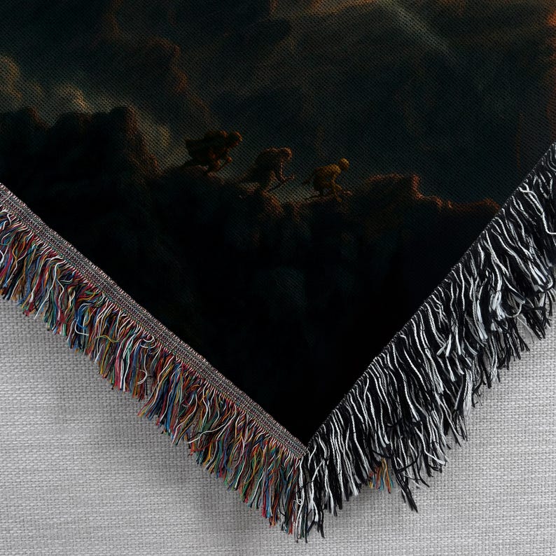 Puede incluir: Una manta oscura con una escena de paisaje dram&aacute;tica y figuras en una monta&ntilde;a. La manta tiene un borde con flecos con una mezcla de colores, incluyendo negro, blanco y un arco&iacute;ris de tonos. La obra de arte representa un entorno sombr&iacute;o y atmosf&eacute;rico.