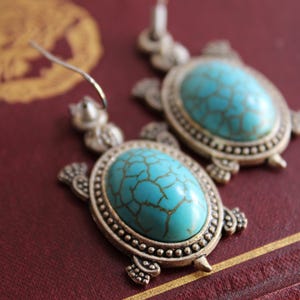 May include: A pair of silver-toned dangle earrings shaped like turtles. Each earring features an oval-shaped, turquoise-colored cabochon with a dark brown webbed pattern set into the shell, suspended from a simple silver-toned metal hook.