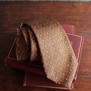 May include: A brown necktie featuring a repeating geometric pattern of dark brown hexagons with orange and light blue accents. The tie is folded and rests on top of two stacked, dark red hardcover books with gold-colored trim on the covers.