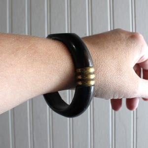 May include: A thick, smooth black bangle bracelet featuring a central decorative element composed of three horizontal, rounded brass-toned metal rings that wrap around the circumference of the dark, solid-colored circular band.
