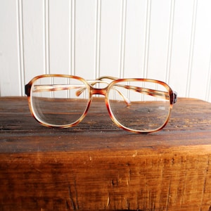 May include: Vintage eyeglasses with a tortoiseshell pattern on the frames. The glasses have a rectangular shape and clear lenses. The frames are a mix of brown, red, and tan colors. The glasses are resting on a wooden surface.