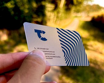 以下が含まれることがあります： 右側に青と白のストライプ模様が施された銀色の名刺。カードには「transparentcard.com」と「parentcard.com」のテキストが印刷されています。電話番号+49 176 6269 7187もカードに印刷されています。