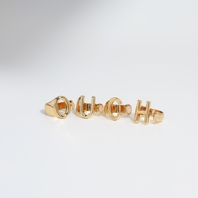 Gold monogram rings spelling out "OUCH" displayed side by side on a white surface, with a sleek minimalist design.