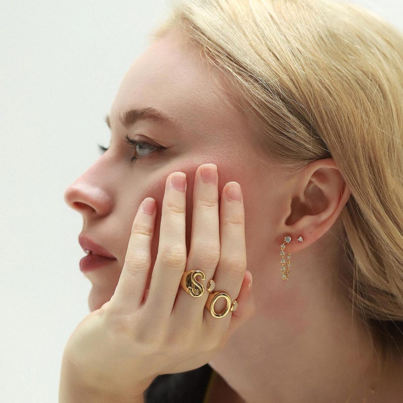 Close-up of a woman wearing gold monogram rings spelling out "SO" paired with dainty gold hoop earrings with CZ accents.