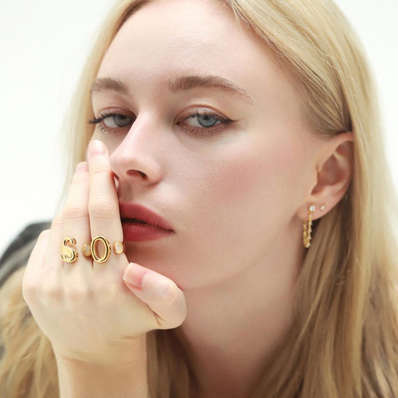 Close-up of a woman wearing gold monogram rings spelling out "SO" paired with dainty gold hoop earrings with CZ accents.
