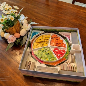 May include: A board game with colorful wooden pieces in a divided circular tray. The pieces are yellow, orange, green, and red. The game is in a box with compartments for cards and other game components. A basket of flowers and eggs is in the background.