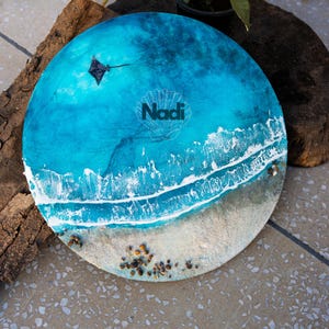 Puede incluir: Obra de arte circular con temática oceánica, con vibrantes olas turquesas y blancas. La obra presenta una zona de playa arenosa con pequeños guijarros y el nombre "Nadi" en un diseño de concha. Se representa una pequeña cometa.
