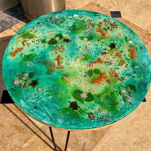 May include: Circular coffee table with a teal resin top depicting a koi pond. The design includes orange fish, lily pads, and shells. The table has a wooden edge and black metal legs. The text "Nadi resin store" is visible on the table.