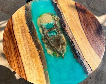 May include: A round wooden table top with a turquoise blue epoxy river running through the center. The river contains a small, weathered boat and sand. The text "Nadkonline Store" is visible in the epoxy.