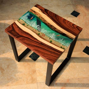 Pode incluir: Mesa lateral quadrada de madeira com um design único de cena oceânica. O tampo da mesa apresenta uma resina epóxi transparente com um design de praia e oceano, incluindo pequenos barcos. A mesa tem uma estrutura de madeira castanha escura e pernas de metal preto.