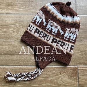 May include: A brown knitted hat with ear flaps and a tassel. The hat features a camel design and the words "PERU" and "ANDEAN ALPACA" in white. The hat is made of a soft, warm material.