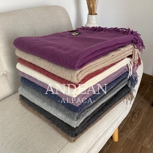 Pode incluir: Uma pilha de cobertores andinos de alpaca coloridos em vários tons, incluindo roxo, bege, vermelho e cinza. Os cobertores estão cuidadosamente dobrados e empilhados, com o texto "ANDEAN - ALPACA-" visível. Os cobertores têm bordas com franjas.