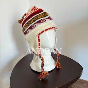 Könnte beinhalten: Eine cremefarbene Strickmütze mit Ohrenklappen, verziert mit horizontalen Bändern aus geometrischen Mustern in Rot, Burgunderrot und Senfgelb. Die Mütze hat geflochtene Quasten an den Ohrenklappen und einen kleinen Bommel, mit dem Text ANDEAN ALPACA.