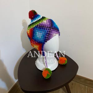 Könnte beinhalten: Ein farbenfroher Andean Alpaka-Hut mit Bommel und Ohrenklappen. Der Hut hat ein Patchwork-Design in Rot-, Orange-, Gelb-, Grün-, Blau- und Lilatönen. Der Text "ANDEAN ALPACA" ist sichtbar. Der Hut wird auf einem weißen Mannequinkopf präsentiert.