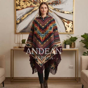 Peut inclure: Un poncho à capuche avec un motif géométrique multicolore dans des tons bordeaux, bleu sarcelle, jaune et crème. Le vêtement présente un design en forme de V, une capuche et une longue frange le long de l'ourlet. Le texte ANDEAN ALPACA est centré.