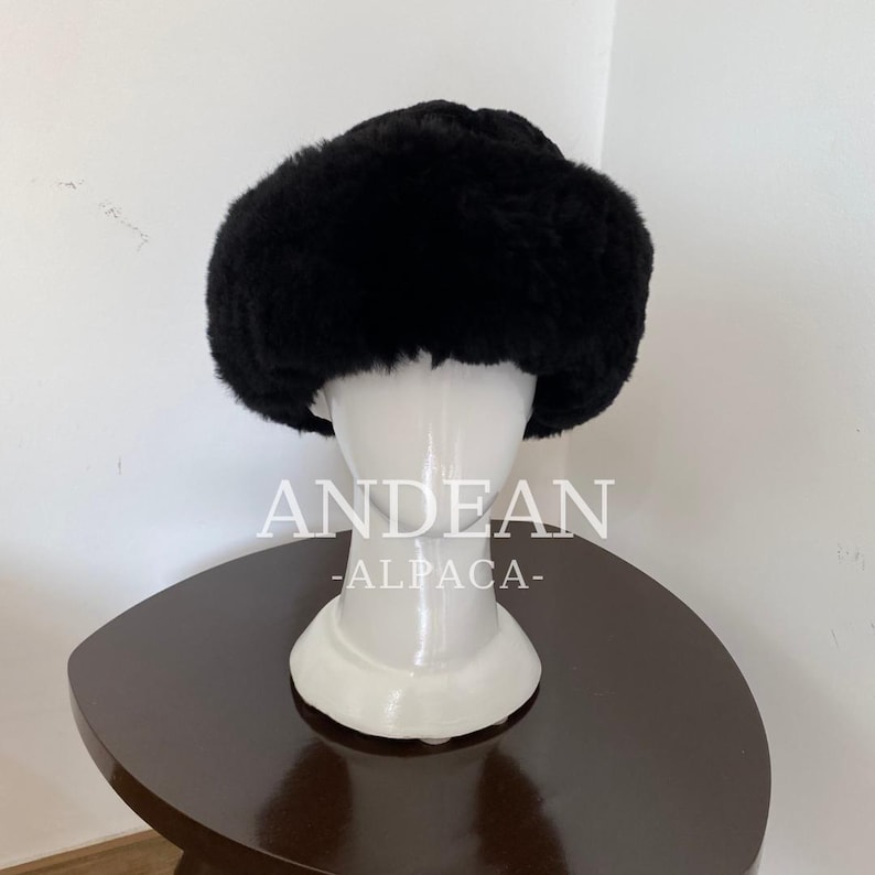 May include: A black, fluffy hat, possibly fur, sits on a white mannequin head. The hat has a rounded shape and looks warm. The text "ANDEAN -ALPACA-" is visible on the mannequin's base. The hat is displayed on a dark brown table.