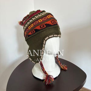 May include: An olive green knit chullo hat featuring geometric patterns in red, orange, and black. The hat includes ear flaps, braided tassels with multi-colored yarn ends, and a pom-pom on top. The text ANDEAN ALPACA is visible on the hat.