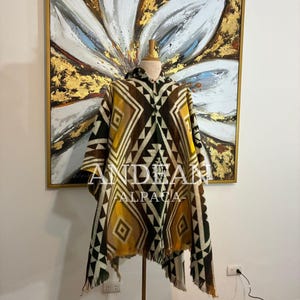 Może przedstawiać: Poncho z kapturem z geometrycznym wzorem w odcieniach brązu, żółci i bieli. Poncho jest prezentowane na manekinie. Tekst "ANDEAN ALPACA" jest widoczny z przodu poncho.
