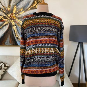 Könnte beinhalten: Ein langärmeliger Pullover mit einem mehrfarbigen Fair-Isle-Muster aus horizontalen Streifen in Orange, Blau, Grün und Schwarz. Das Strickkleidungsstück zeigt den Text ANDEAN ALPACA auf der Brust und hat einen schwarzen, gerippten Rundhalsausschnitt.