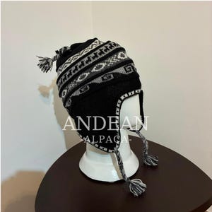 May include: A black and grey knit earflap hat featuring a geometric pattern and braided tassels. The hat displays the text ANDEAN ALPACA in white lettering across the front. The design includes a solid black base with grey accents.
