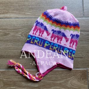 Puede incluir: Gorro de punto rosa con un colorido diseño peruano. El gorro presenta un patrón de arcoíris, llamas moradas y las palabras "PERU PERU" en azul y amarillo. El gorro también tiene el texto "ANDEAN - ALPACA".