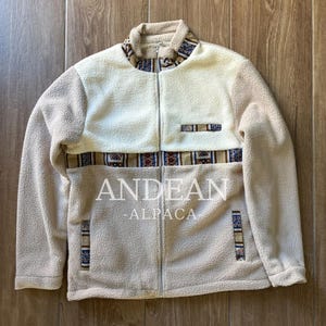 Könnte beinhalten: Eine beigefarbene und cremefarbene Sherpa-Fleece-Jacke mit Reißverschluss und einem horizontalen Band mit geometrischem Muster in Blau, Braun und Weiß. Die Jacke hat eine kleine Brusttasche und zwei untere Taschen mit passendem Besatz und dem Text ANDEAN ALPACA.