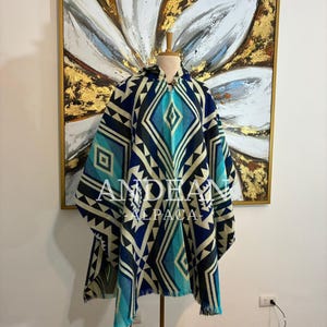 Op de afbeelding: Een blauwe, turquoise en crèmekleurige poncho met een geometrisch ruitpatroon. De poncho heeft een capuchon en franjes aan de onderkant. De tekst "ANDEAN ALPACA" is zichtbaar op de voorkant.