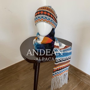 May include: A knitted beanie and matching scarf set featuring a multi-coloured geometric pattern in shades of orange, blue, white, and black. The scarf includes a fringed edge and the text ANDEAN-ALPACA is visible across the centre of the image.