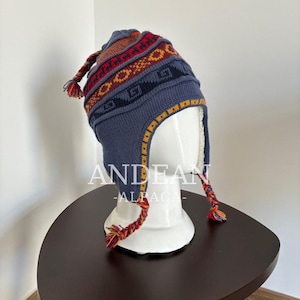 Könnte beinhalten: Eine blaue Strickmütze mit Ohrenklappen und einem geometrischen Muster in Rot, Orange und Gelb. Die Mütze hat eine geflochtene Quaste oben und zwei geflochtene Bänder an den Ohrenklappen mit bunten Enden. Das Innere ist mit weißem Fleece gefüttert.