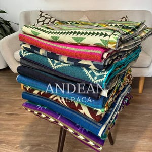 Puede incluir: Una pila de mantas tejidas dobladas con varios patrones geométricos y colores vibrantes que incluyen verde, rosa, azul marino, verde azulado y morado. El texto ANDEAN ALPACA está impreso en blanco en la parte frontal de la pila de textiles.
