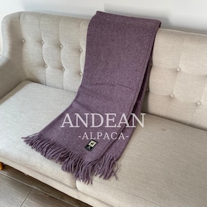 May include: A lavender-colored Andean alpaca scarf with fringe. The scarf is draped over a light gray sofa. The text "ANDEAN -ALPACA-" is printed on the scarf. A small tag with a llama logo is visible.