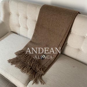 May include: A brown Andean Alpaca throw blanket with fringe detail, draped over a beige sofa. The blanket features the text "ANDEAN -ALFACA-" in white. The throw is a solid color and appears soft and warm.
