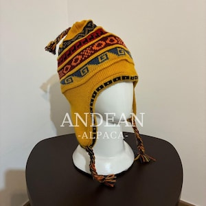 Könnte beinhalten: Eine senfgelbe Strickmütze mit Ohrenklappen, geflochtenen Quasten und einem mehrfarbigen geometrischen Muster in Rot, Orange und Blau. Die Mütze zeigt den Text ANDEAN-ALPACA in weißen Buchstaben auf der Vorderseite des gestrickten Stoffes.