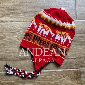 Può includere: Cappello andino rosso in alpaca con disegno di cammello e paraorecchie. Il cappello presenta la scritta "PERU" e "ANDEAN ALPACA". Il cappello ha un pompon bianco sulla parte superiore e un nappa intrecciata.