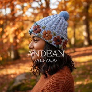Könnte beinhalten: Hellblaue Strickmütze mit Zopfmuster und passendem Bommel. Die Mütze ist mit gestickten Blumen in Orange-, Braun- und Grüntönen verziert. Der Text "ANDEAN -ALPACA-" ist auf dem Bild zu sehen.