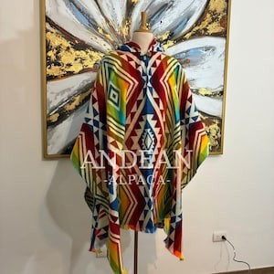 Könnte beinhalten: Ein farbenfrohes Anden-Alpaka-Poncho mit geometrischem Muster in Rot, Blau, Gelb und Grün. Der Poncho hat eine Kapuze und Fransen. Der Text "ANDEAN ALPACA" ist auf der Vorderseite zu sehen.