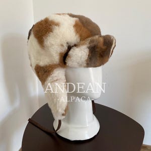 Op de afbeelding: Een trappermuts met oorflappen, met een patroon van bruine en witte vlekken. De muts is gemaakt van een zacht, pluizig materiaal en heeft een bruin touwtje. De tekst "ANDEAN ALPACA" is zichtbaar.