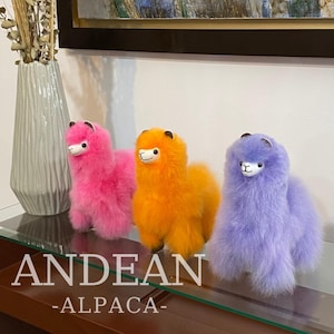 May include: Three plush alpaca toys in pink, orange, and purple. The toys are sitting on a wooden surface with the text "ANDEAN -ALPACA- ".