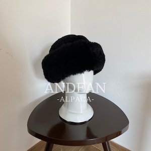 May include: A black fur hat with a wide brim, displayed on a white mannequin head. The hat is fluffy and appears warm. The mannequin is on a dark brown, three-legged table. The text "ANDEAN -ALPACA-" is visible.
