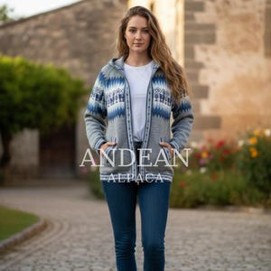 Peut inclure: Un pull à capuche gris zippé avec des motifs géométriques bleus et blancs. Le pull a deux poches avant et le texte "ANDEAN ALPACA" imprimé sur le devant. La personne sur l'image porte un jean bleu et un t-shirt blanc.