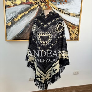 Peut inclure: Un poncho à capuche avec un motif géométrique noir, gris et crème, doté d'un bouton en bois au niveau de l'encolure. Le vêtement présente un ourlet à franges et affiche le texte ANDEAN ALPACA au centre du motif du tissu.