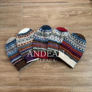 May include: Five colourful knit beanies with intricate geometric patterns and the text "ANDEAN ALPACA". The hats feature a variety of colours, including blue, red, brown, and white, and are displayed on a wooden surface.