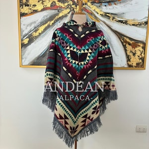 Op de afbeelding: Een poncho met capuchon en een geometrisch patroon in tinten blauwgroen, bordeauxrood, crème en grijs. Het kledingstuk heeft een enkele knoopsluiting bij de halslijn, een puntige zoom en grijze franjes langs de randen. De tekst ANDEAN ALPACA is zichtbaar.