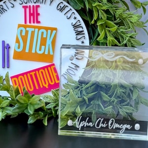 May include: Clear acrylic magnetic frames with engraved designs, featuring the text "Alpha Chi Omega." The frames are offered in 4" x 4" and 4" x 6" sizes. The image also includes a logo for "The Stick Boutique."